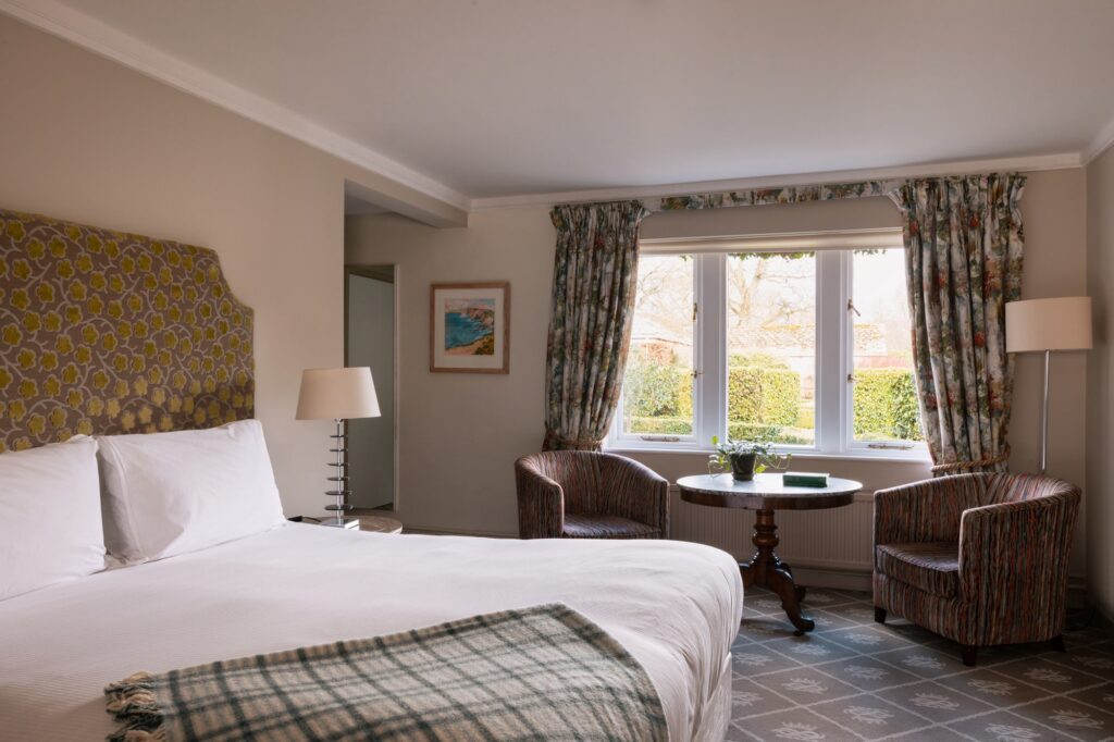 A beige room with two chairs in front of a large window looking out towards the garden, with check blue blanket on the bed.