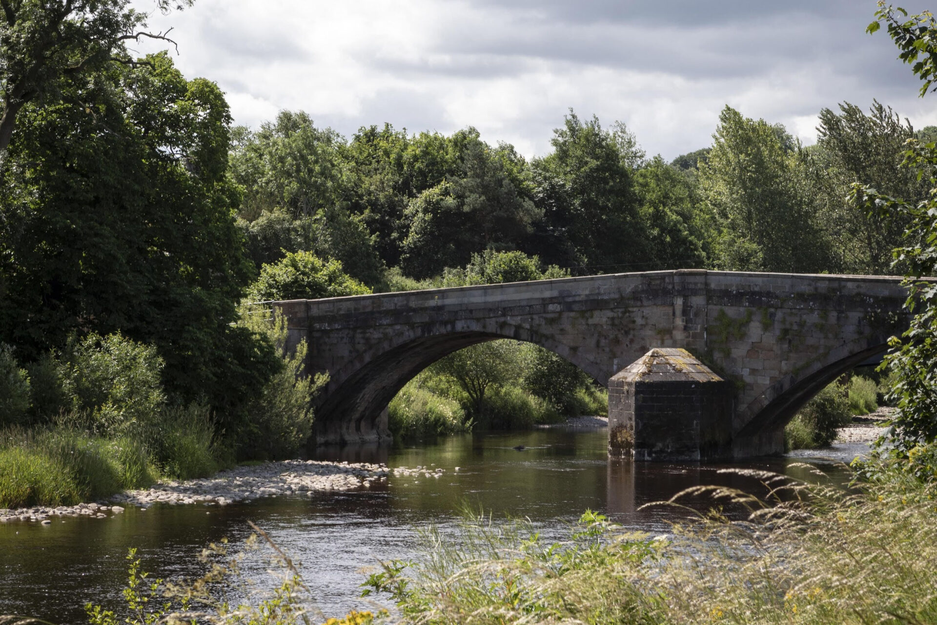 A large stone bridge goes over the river with trees surrounding it.