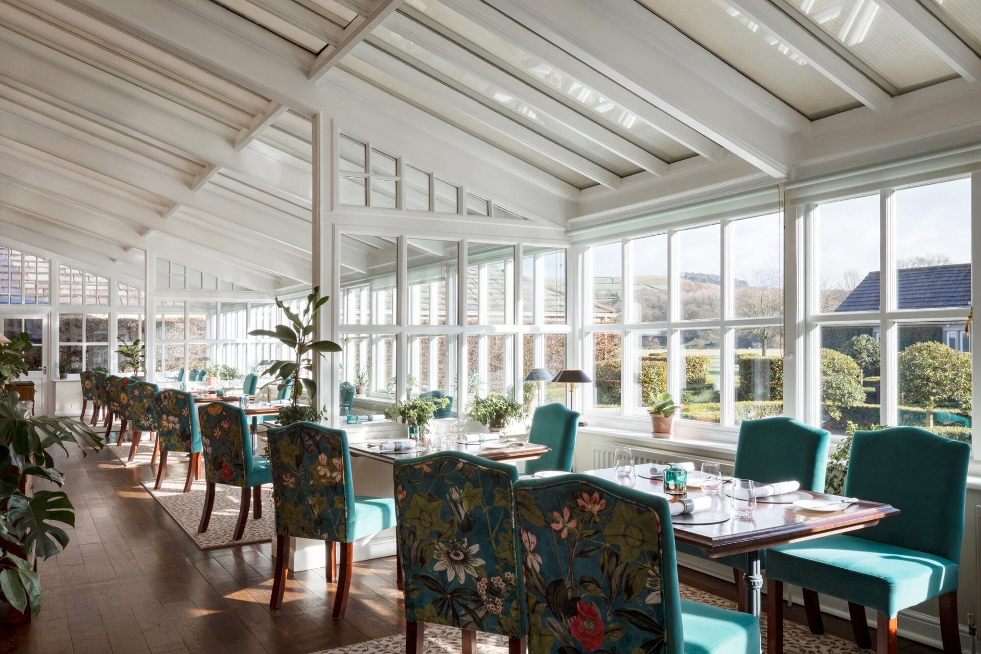 A large conservatory with lots of plants and big windows letting the light in, with green floral chairs at tables.