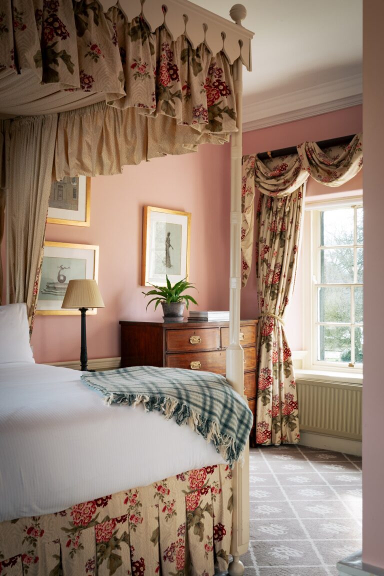 A large fourposter bedroom with pink walls and floral curtains, with antique furnishings.