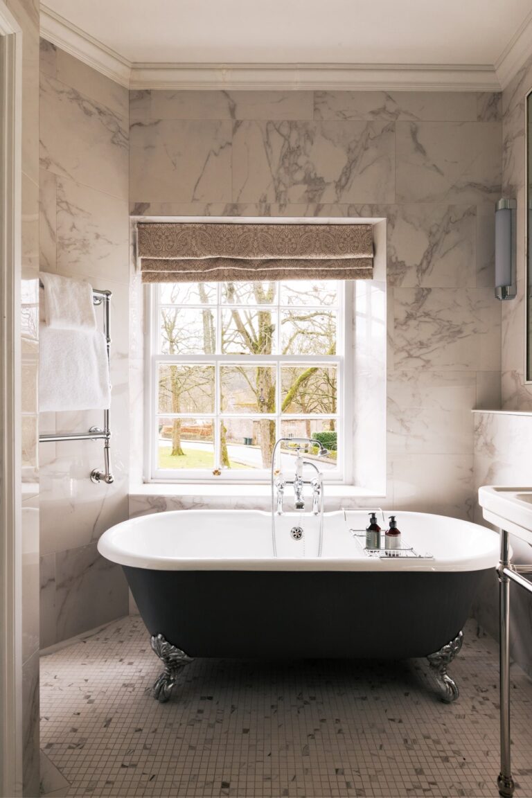A large roll top bath in a marble bedroom, with view out of the large window.