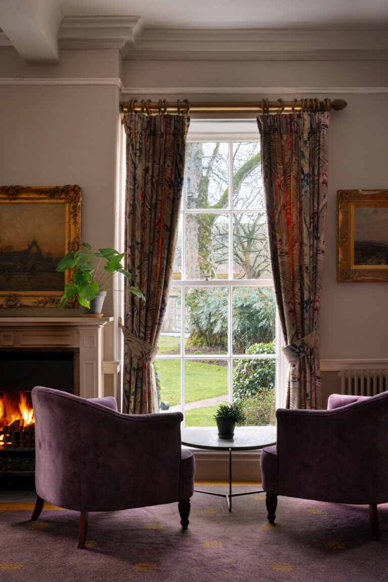 A cosy fire in the lounge, two purple armchairs look out a large window to the garden view.