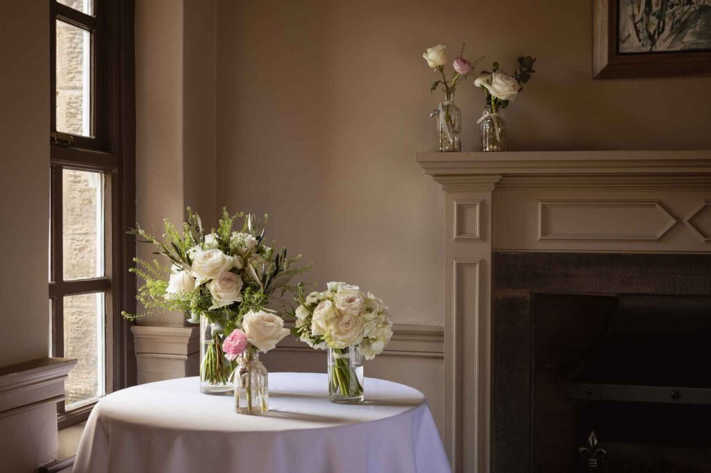 Set of three flowers on a table with flowers on the mantlepiece behind