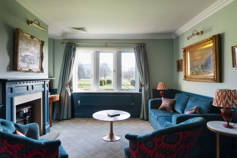 A large suite room with blue walls and floral details, with a sitting room, antique furniture and large windows with views of the rolling hills.