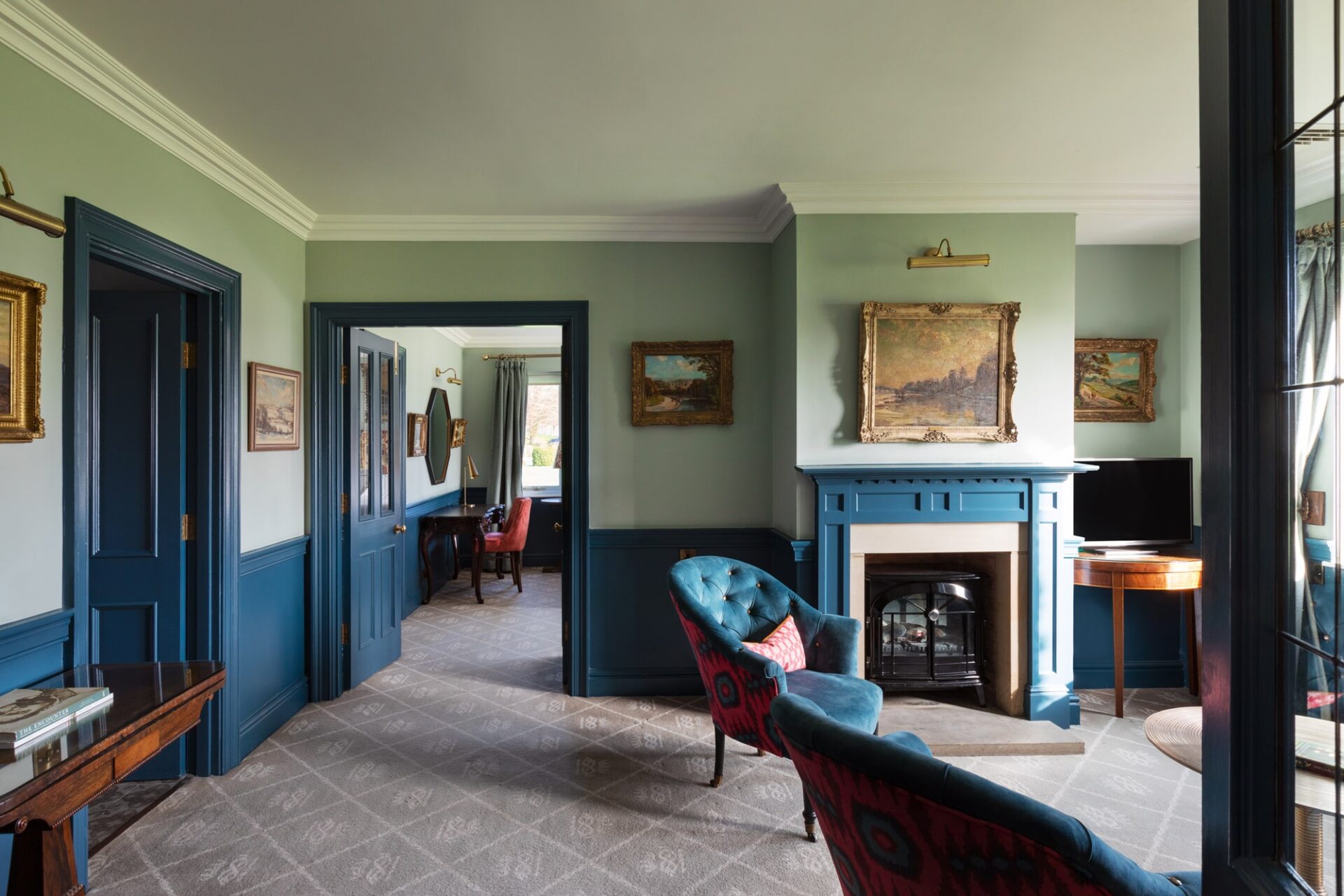 A large suite room with blue walls and floral details, with a sitting room, antique furniture and large windows with views of the rolling hills.