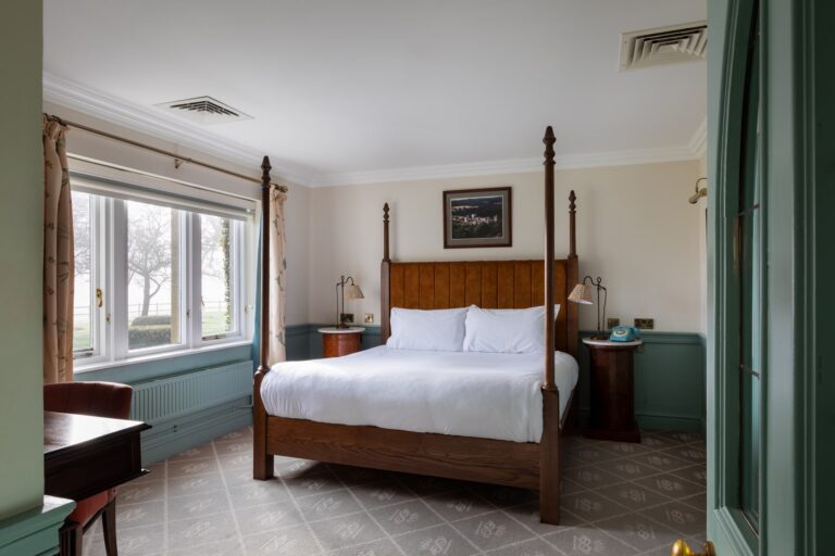 A large suite room with pale green walls and pink details, with a sitting room, antique furniture and large windows with views of the rolling hills.