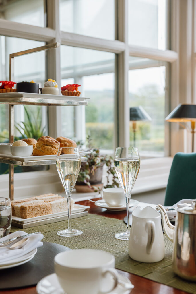 A luxurious afternoon tea stand in The Garden Room filled with sandwiches, sweet treats and scones. A large pot of tea also lays on the table. Two large glasses of Prosecco and a pot of tea lays on the table.